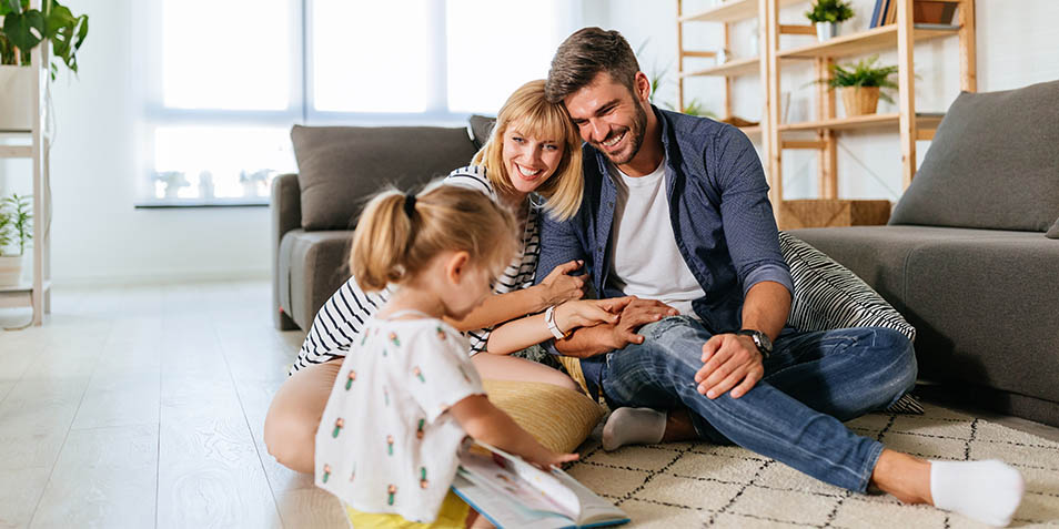 Mother and father sit on the carpet and smile at their playing child (Photo)