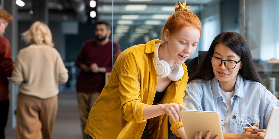 A red-haired employee shows a dark-haired colleague something on her tablet. (Photo)