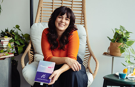 Woman is sitting in a chair smiling. Next to her are books and plants (Photo)